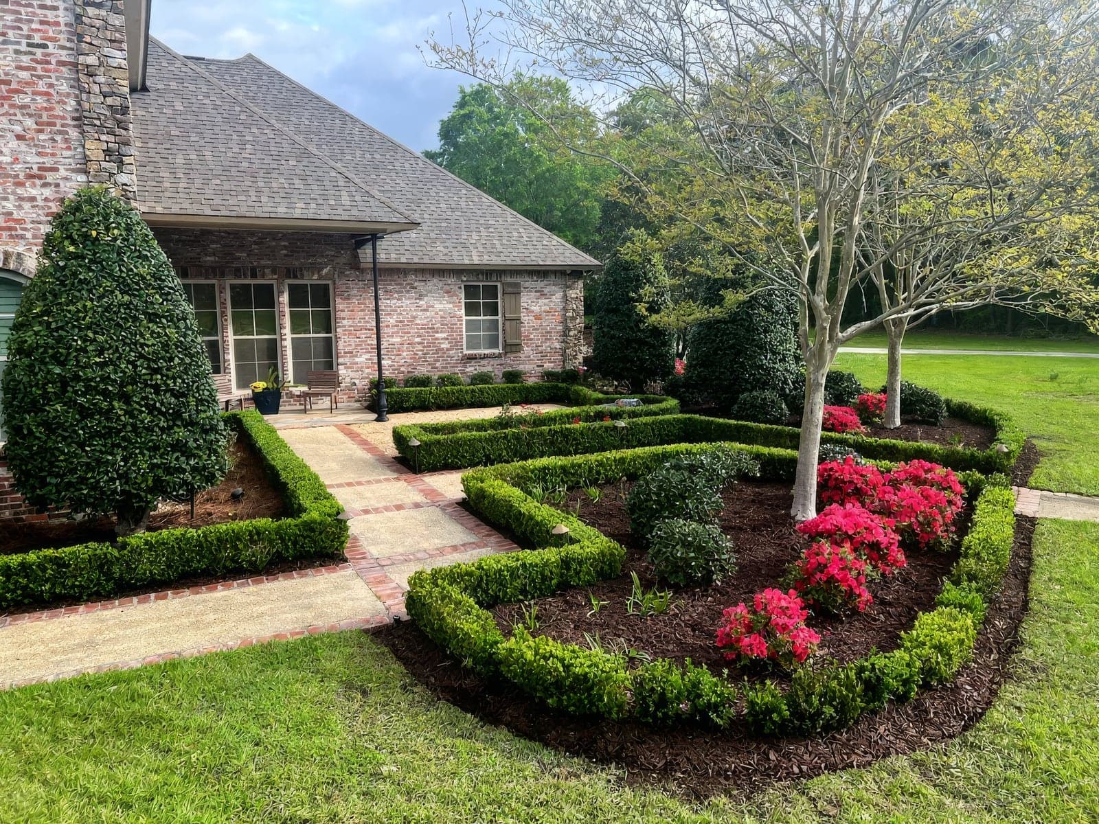 Professionally landscaped front yard featuring manicured boxwood hedges, mulched flower beds with vibrant pink azaleas, and mature trees framing a brick home