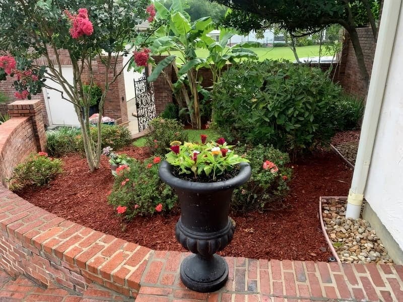 A well-manicured garden bed with mulch, flowering plants, shrubs, and a black planter urn, bordered by a brick wall along a home's entrance walkway