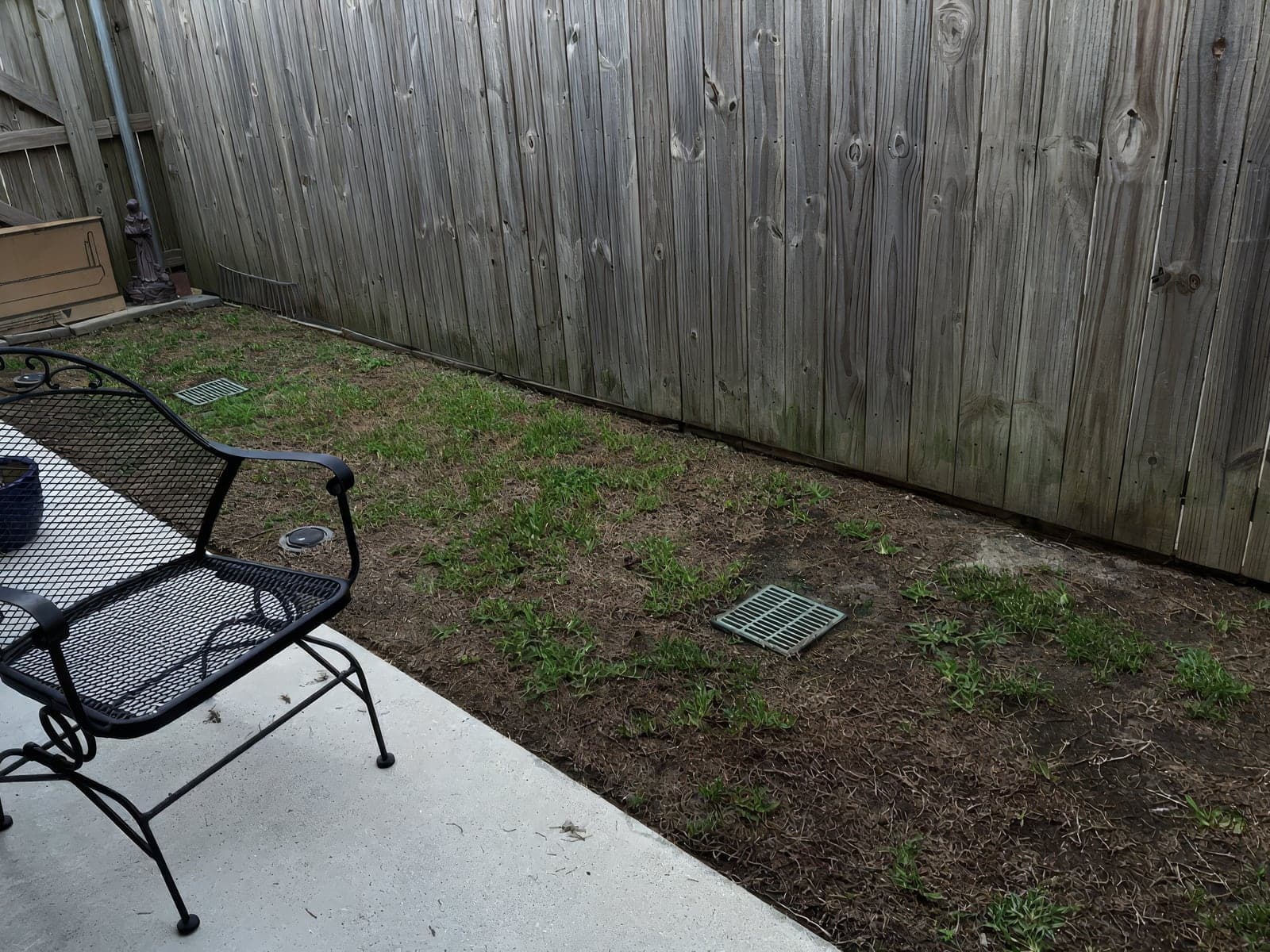 A patchy backyard lawn with bare and dead grass patches alongside a wooden privacy fence, with patio furniture in the foreground
