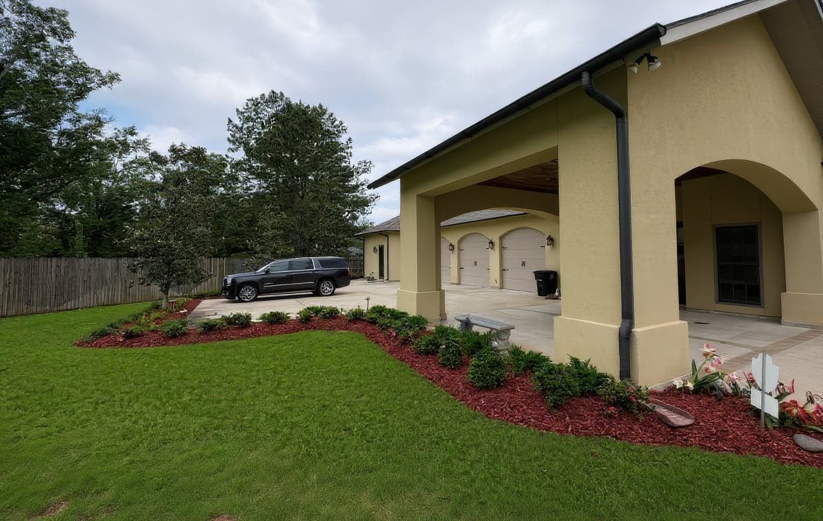 A landscaped residential property with manicured lawn, fresh mulch beds, shrubs, and flowers alongside a stucco home with attached garage and SUV in driveway