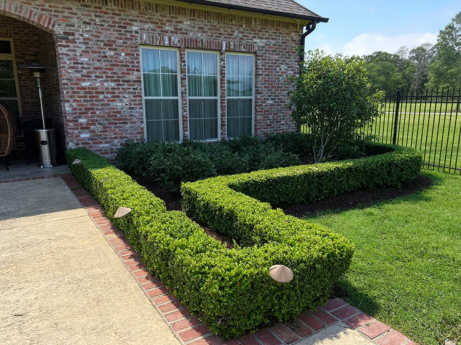 Neatly manicured boxwood hedges and landscaped garden bed alongside a brick house, showcasing professional lawn and landscape maintenance