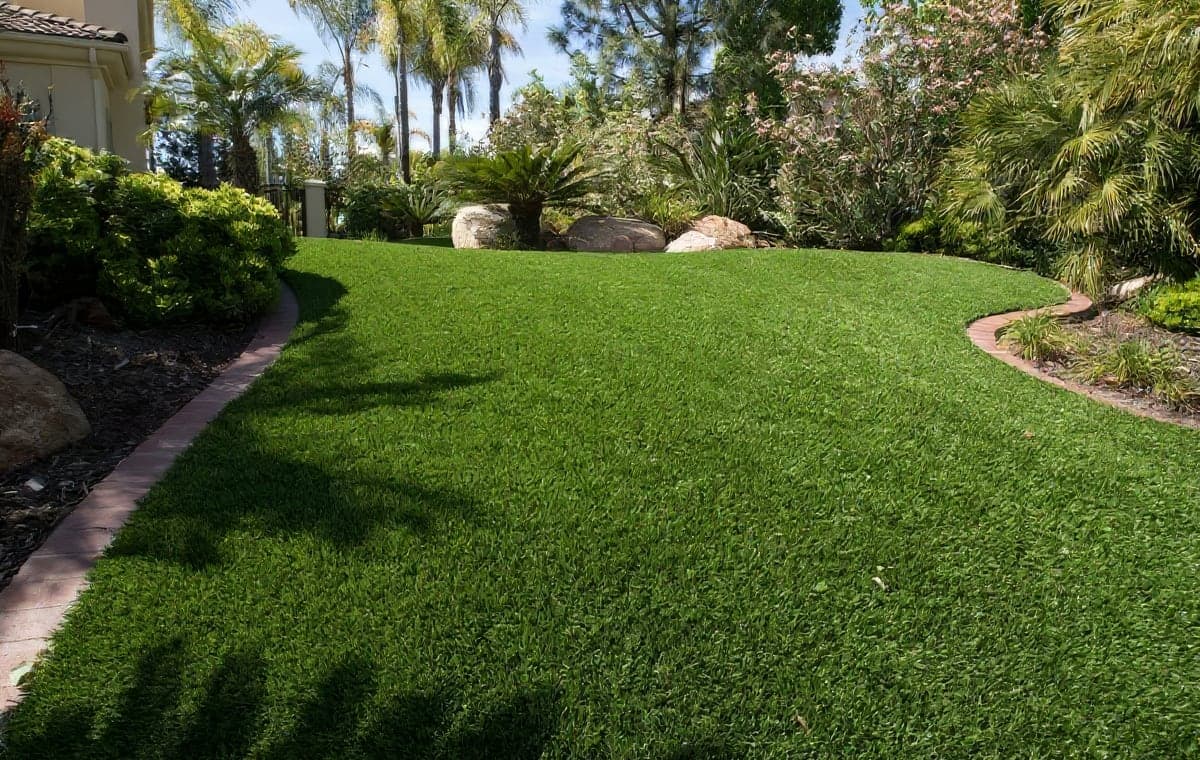 A lush backyard landscape featuring a manicured artificial or well-kept lawn bordered by brick edging, tropical palm trees, and flowering shrubs