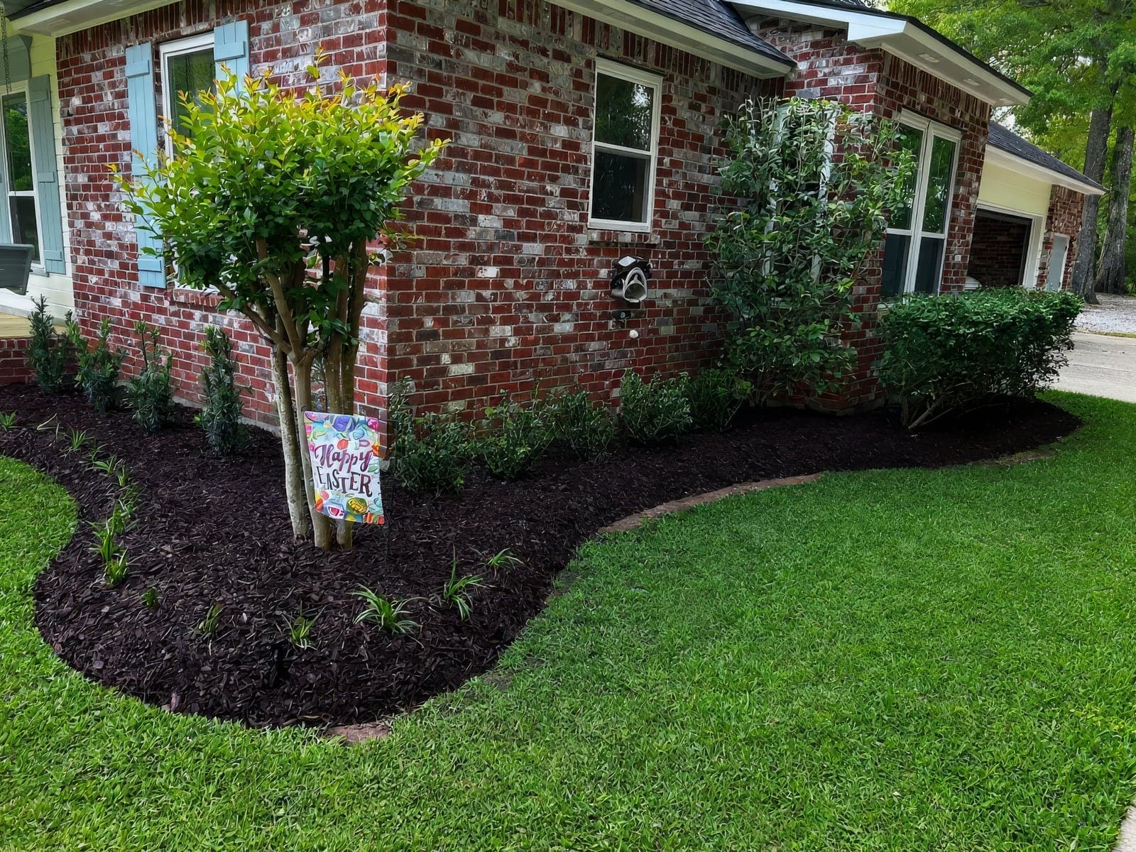 A freshly landscaped garden bed with mulch, new shrubs, and a small ornamental tree along the brick exterior of a home, decorated with a Happy Easter sign