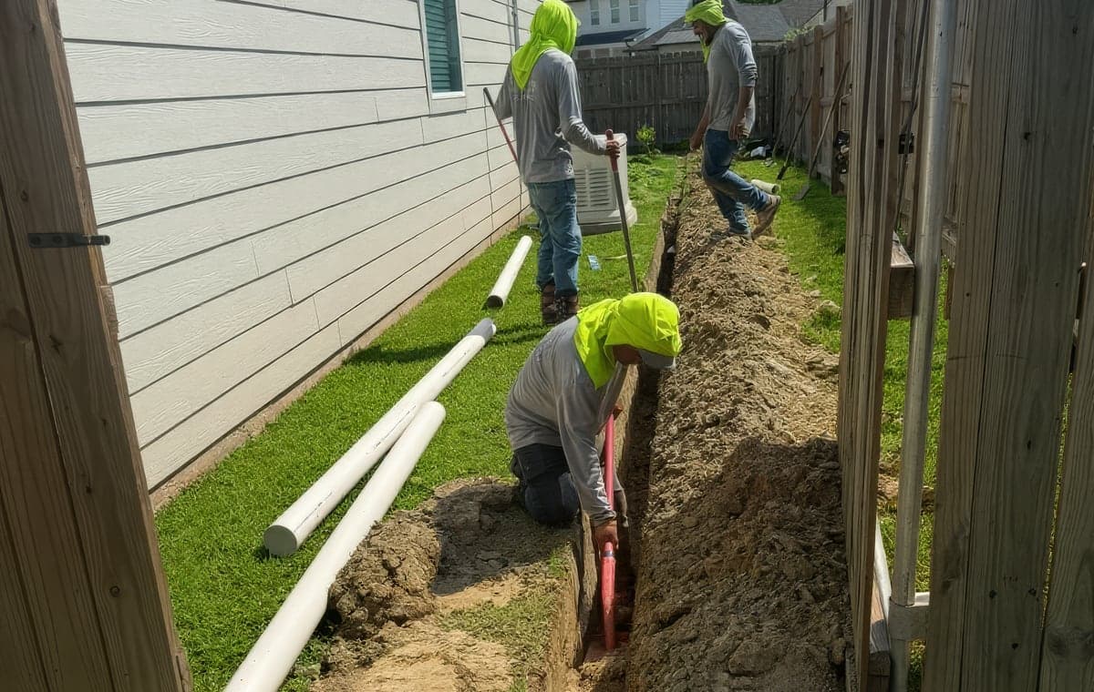 Three crew members installing PVC drainage pipe in a trench along a side yard