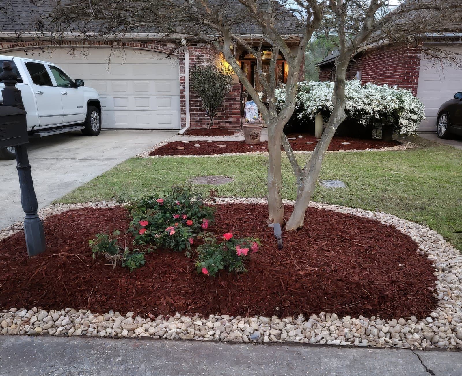 A landscaped residential front yard featuring a mulched flower bed with rock borders, rose bushes, and a mature crepe myrtle tree in front of a brick home