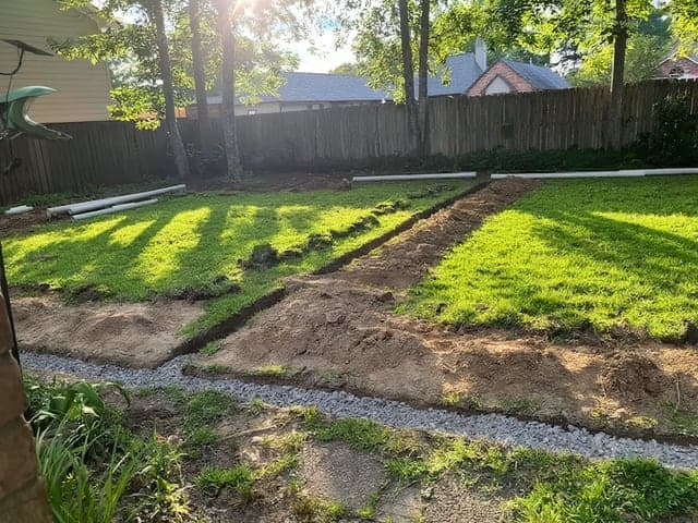 A residential backyard mid-landscaping project with newly laid sod strips, fresh soil trenches, and a gravel drainage channel being installed