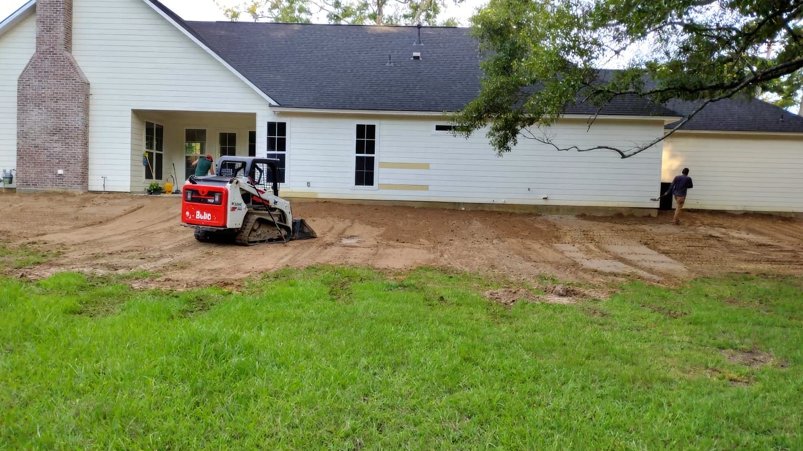 A skid steer grading a backyard to bare soil behind a white home before new sod
