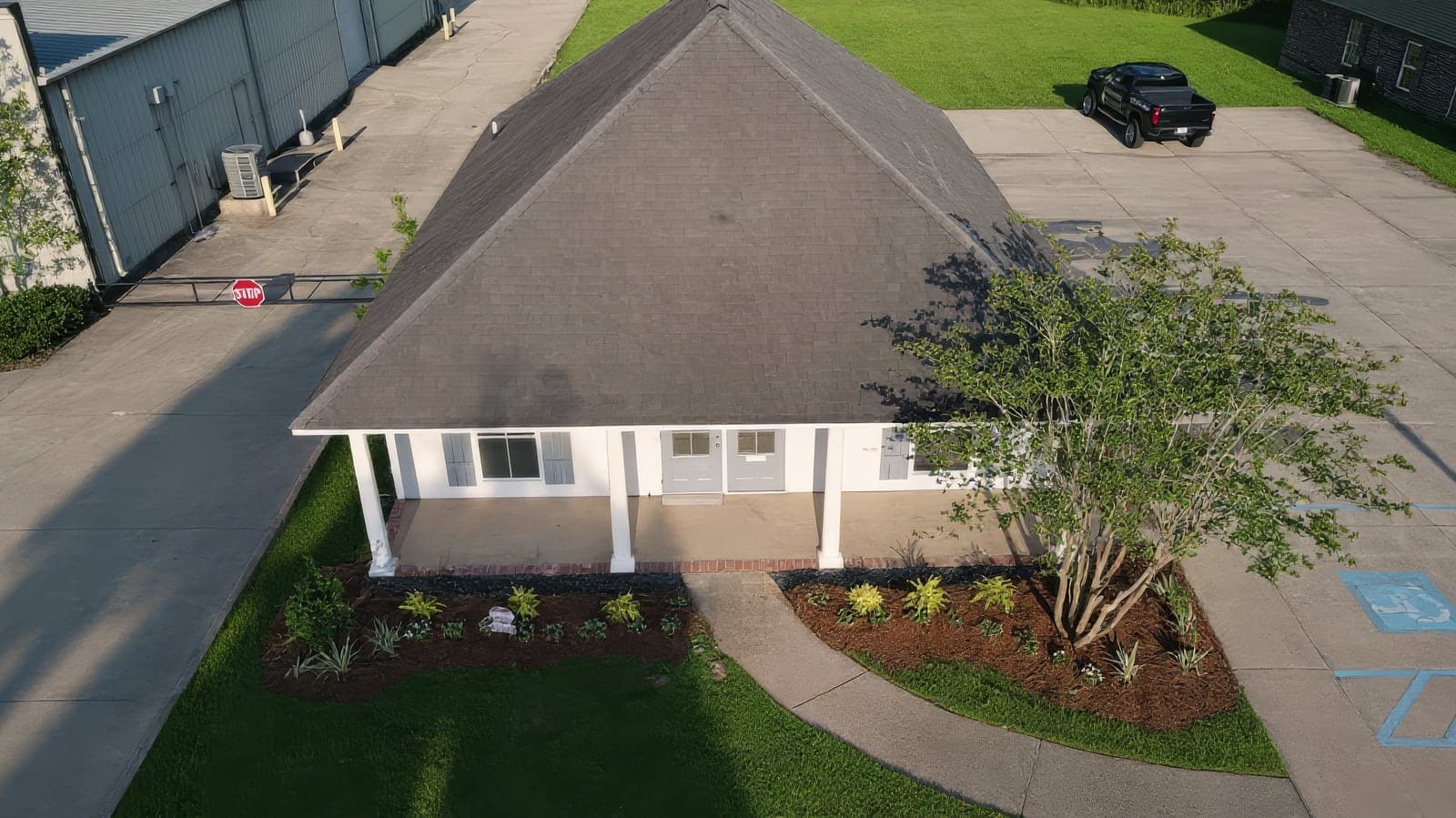 Aerial view of a small commercial building with a white front porch and manicured landscaping beds, surrounded by a paved parking lot