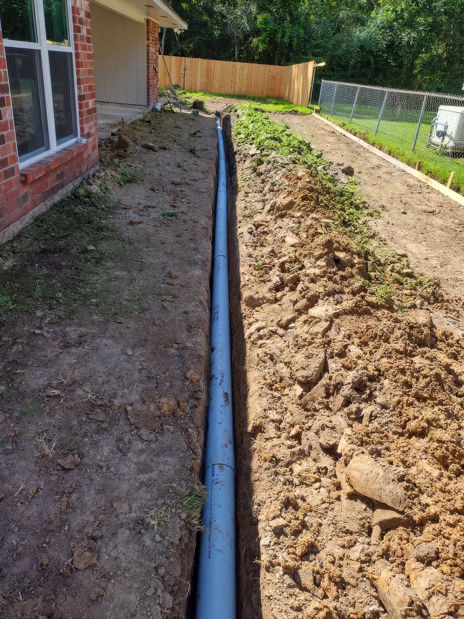 A gray PVC drainage pipe laid in a trench beside a brick home