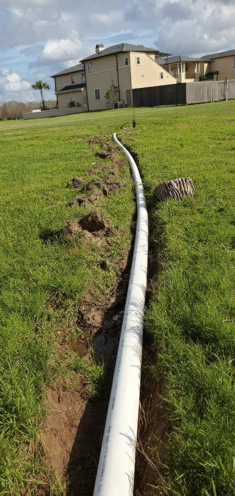 A white PVC pipe laid alongside a freshly dug trench in a grassy lawn, part of an irrigation or drainage installation, with a large tan two-story home in the background
