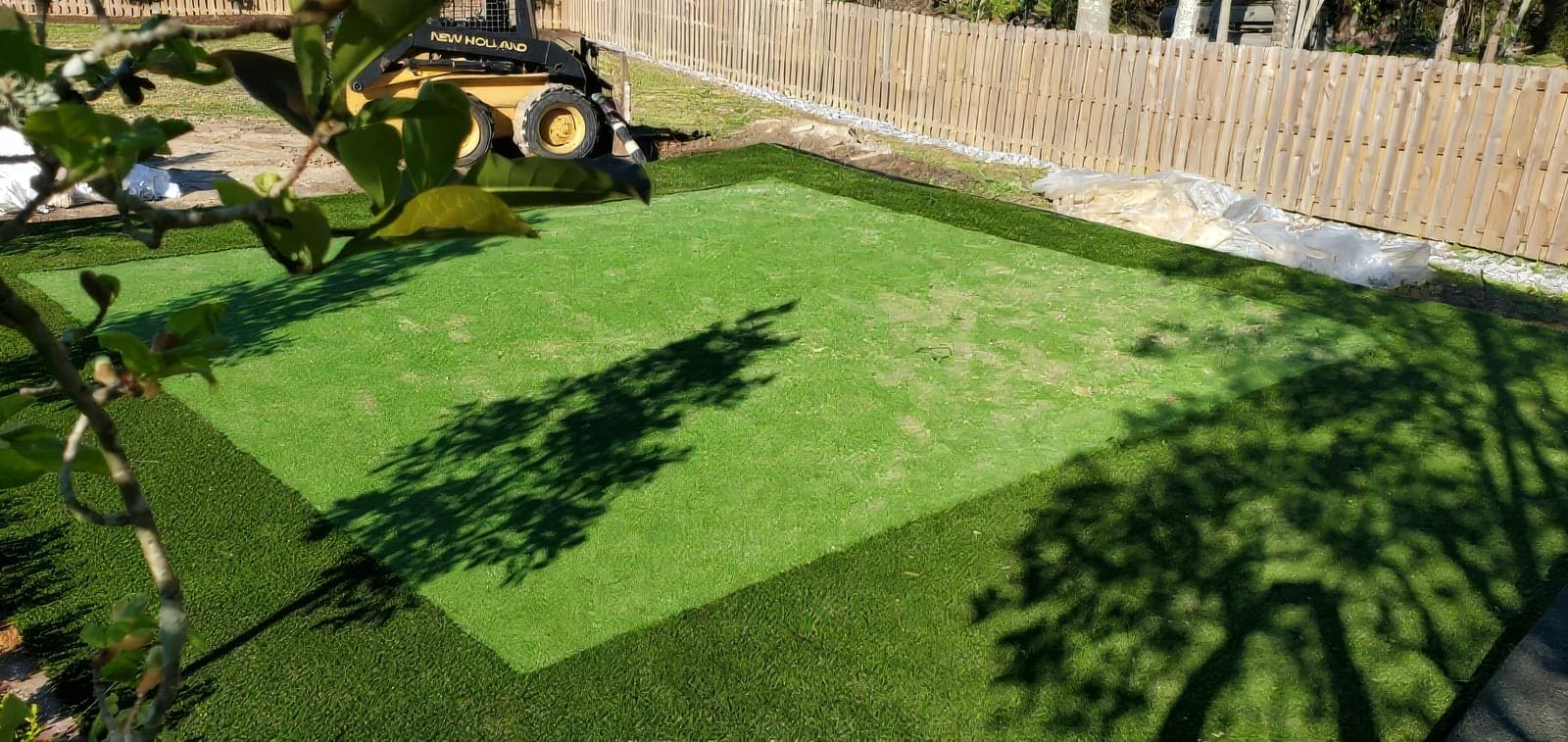 A freshly installed artificial turf area in a fenced backyard, with a lighter green center panel bordered by darker green turf. A New Holland skid steer and leftover installation materials are visible in the background