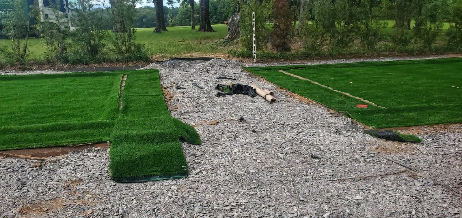 Artificial turf installation in progress on a gravel base, with two sections of green turf laid on either side of an exposed gravel pathway. A branded landscaping truck and mature trees are visible in the background