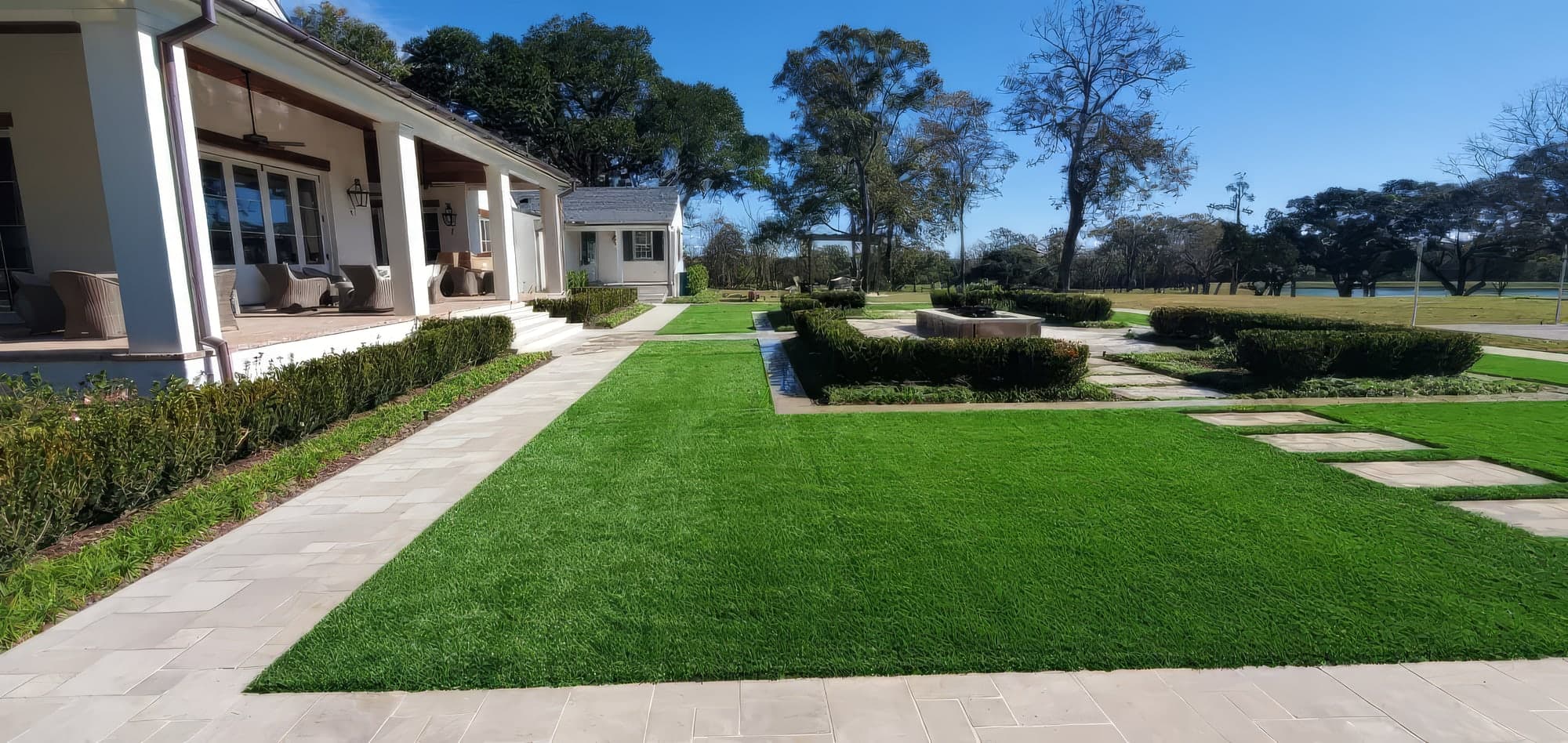 A freshly installed artificial turf lawn framed by clean stone paver walkways and manicured boxwood hedges beside an elegant white home with a covered patio. A formal garden with a fire feature and mature oak trees exten