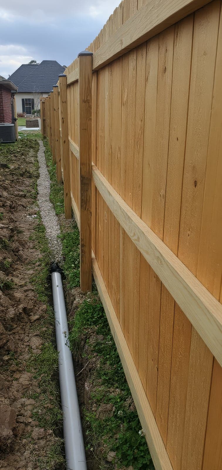 A newly built wooden privacy fence runs alongside a freshly dug trench containing a gray PVC drainage pipe and a gravel-lined channel, installed between a brick home and the fence line. The photo shows in-progress yard d