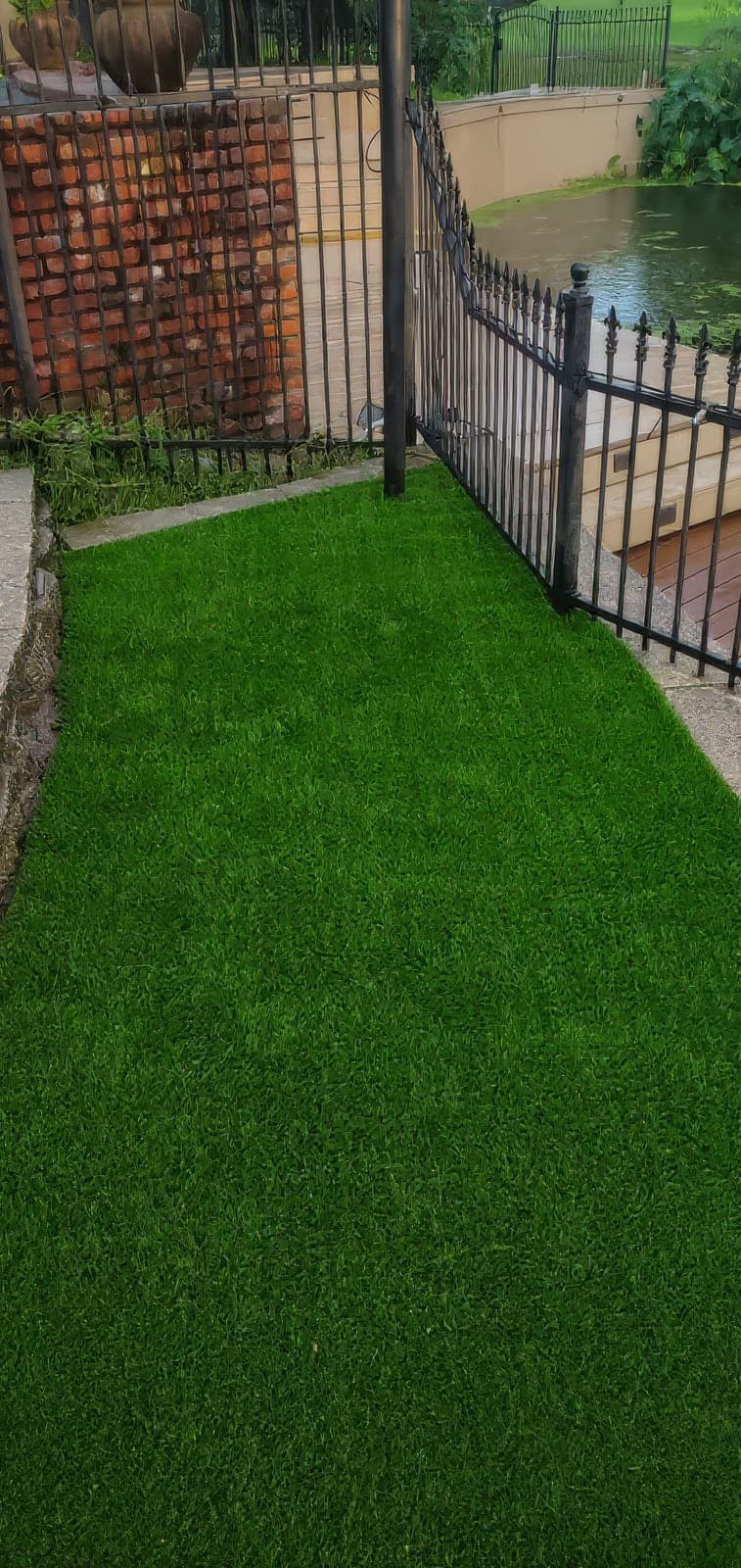 Freshly installed lush green artificial turf in a narrow yard area bordered by a black wrought-iron fence, brick wall, and stone edging, with a pond and wooden steps in the background