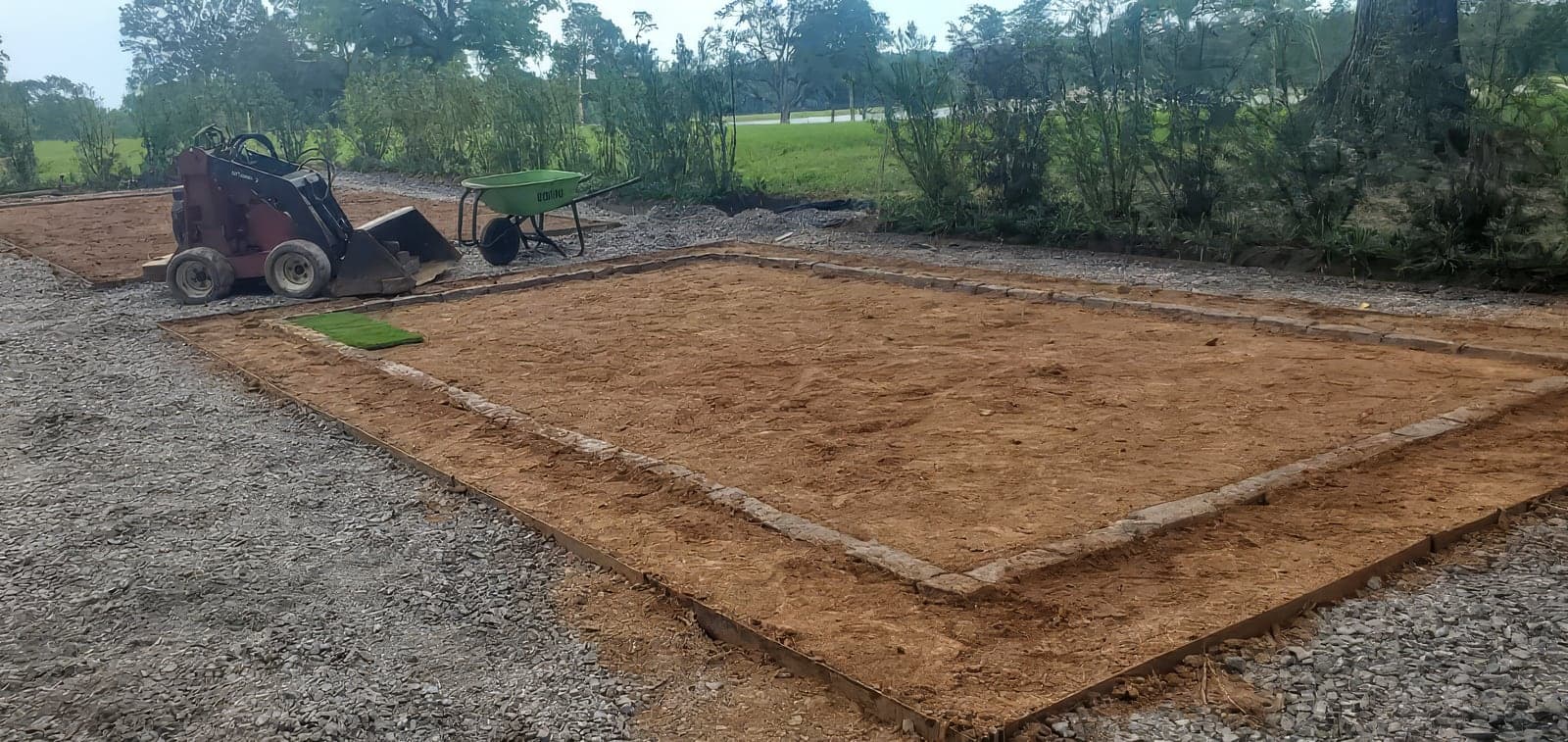 A landscaping job site showing a graded and prepared soil bed framed with steel edging and stone borders, with a small sod sample laid in the corner. A compact skid steer loader and a green wheelbarrow sit nearby, with g