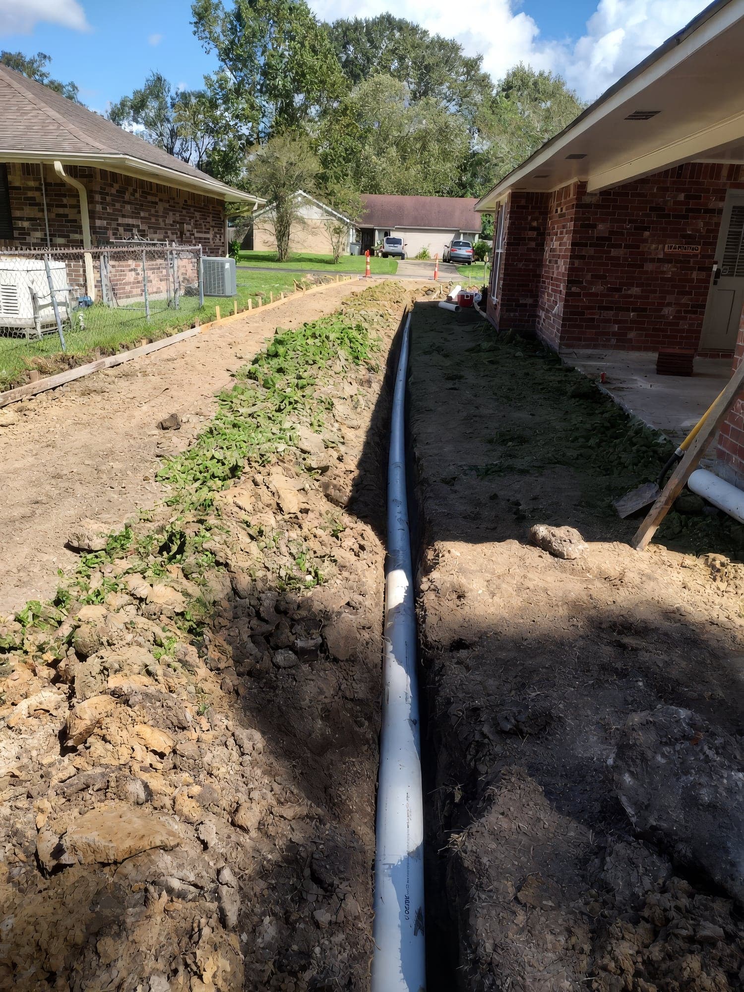 A six-inch PVC drainage pipe laid in a long open trench between two brick homes