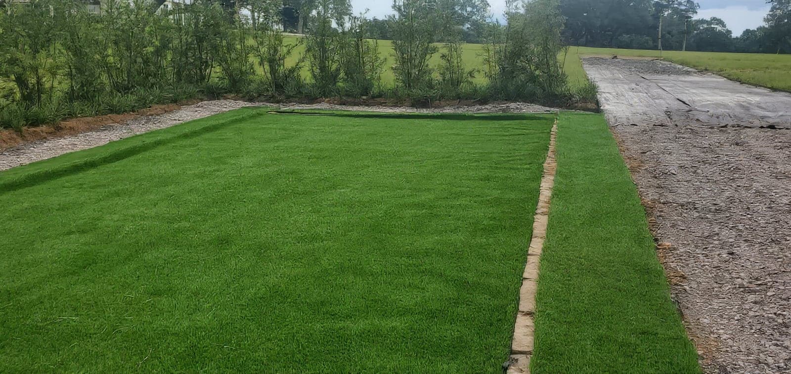Freshly installed lush green lawn section with a neat stone edging border, surrounded by gravel and landscaped shrubs on a spacious property. The finished turf shows clean lines and professional installation
