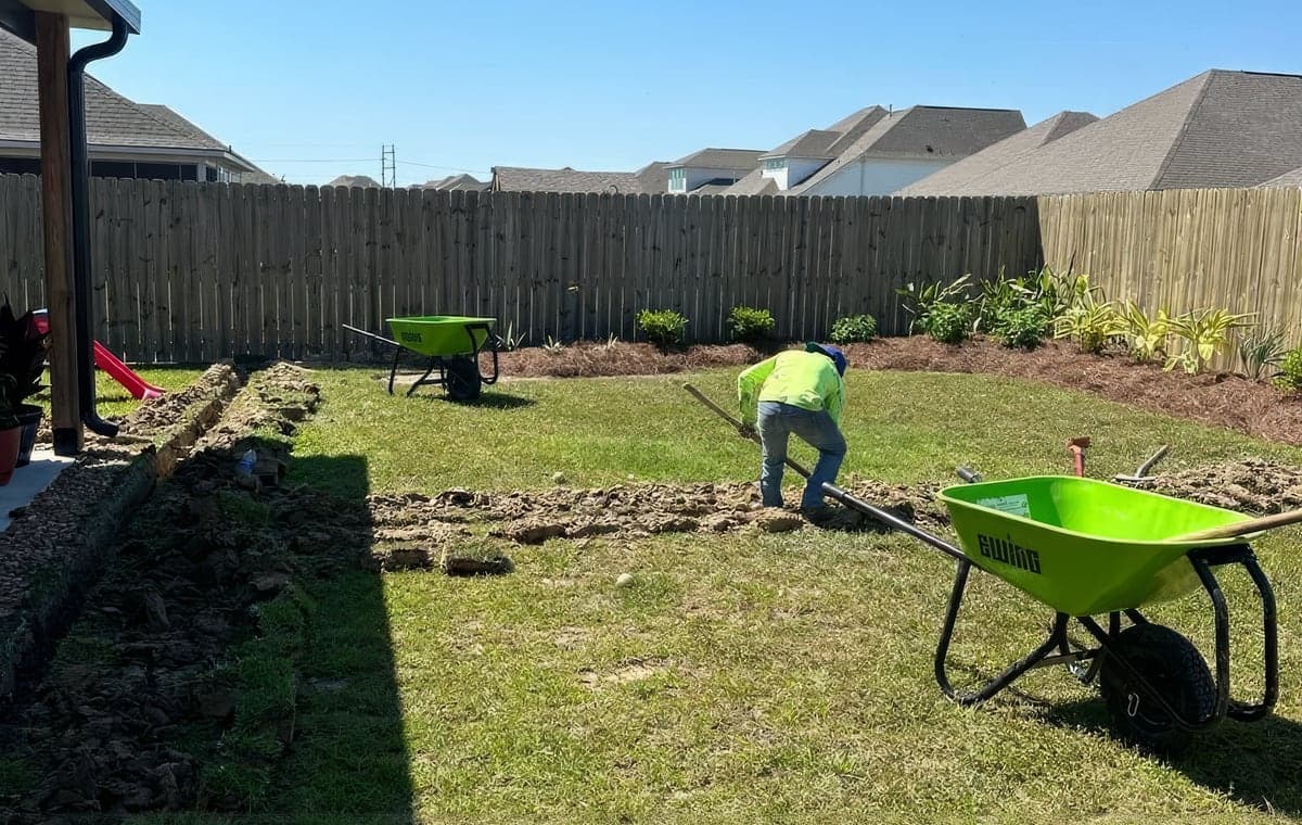 A Hernandez Lawnscape crew member in a high-visibility shirt preparing a bed by hand in a Baton Rouge backyard