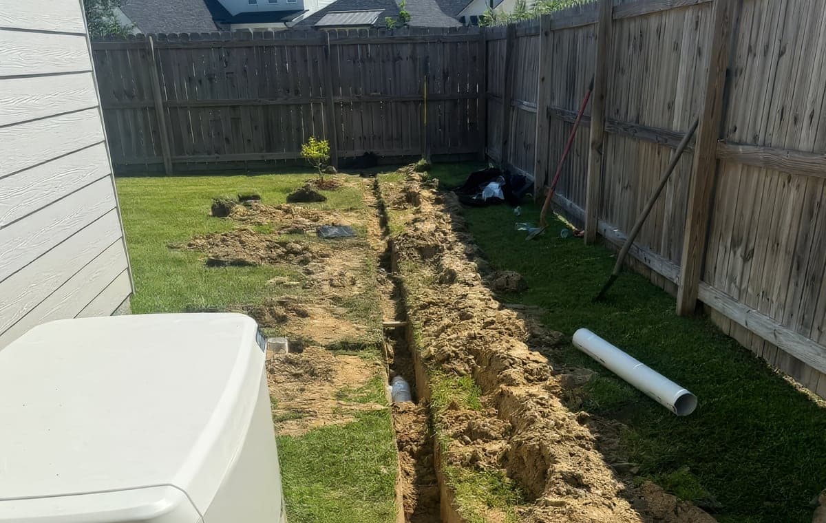 An open trench dug along a wooden privacy fence in a residential backyard with PVC drainage pipe already laid in the trench and another length of white pipe lying on the grass. Shovels and tools lean against the fence, s