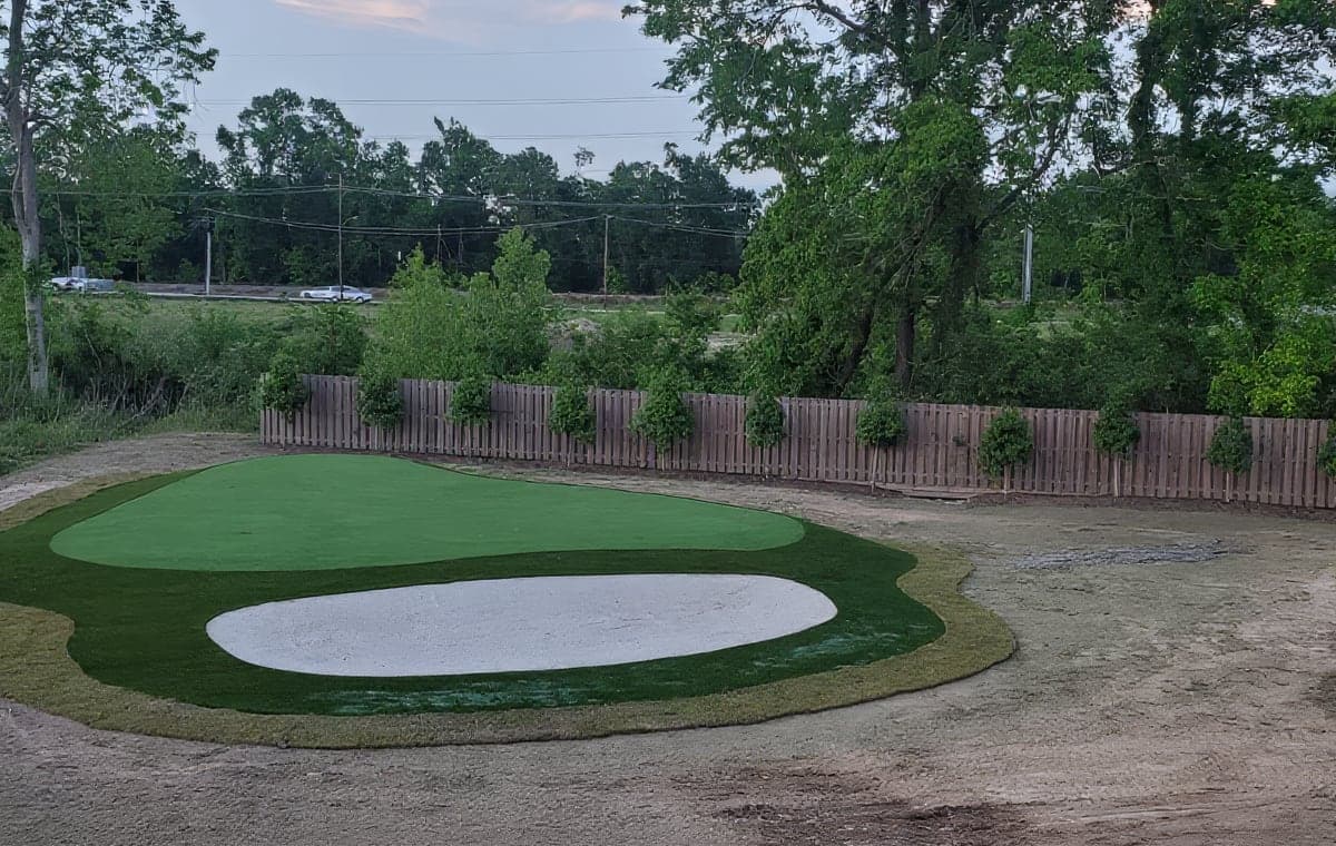 A newly installed backyard artificial turf putting green with a sand-colored bunker feature and a tidy sod border, set along a wooden privacy fence lined with young evergreen shrubs. The surrounding yard is still bare di