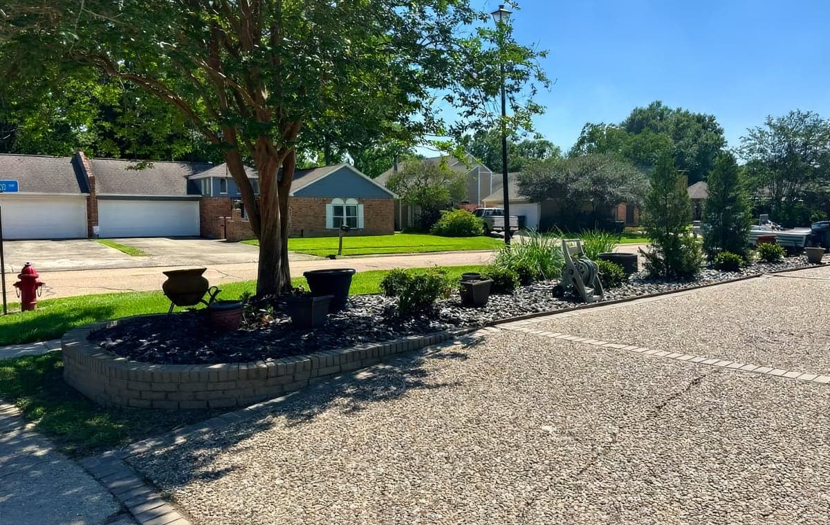 A curved brick-bordered bed with dark river rock, ornamental grasses and a crape myrtle