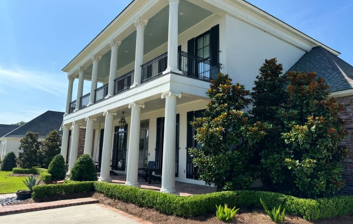 A two-story white stucco home with tall Corinthian-style columns, black shutters, and wrought-iron balcony railings. The front is framed by healthy magnolia trees, neatly trimmed boxwood hedges, and fresh pine straw mulc