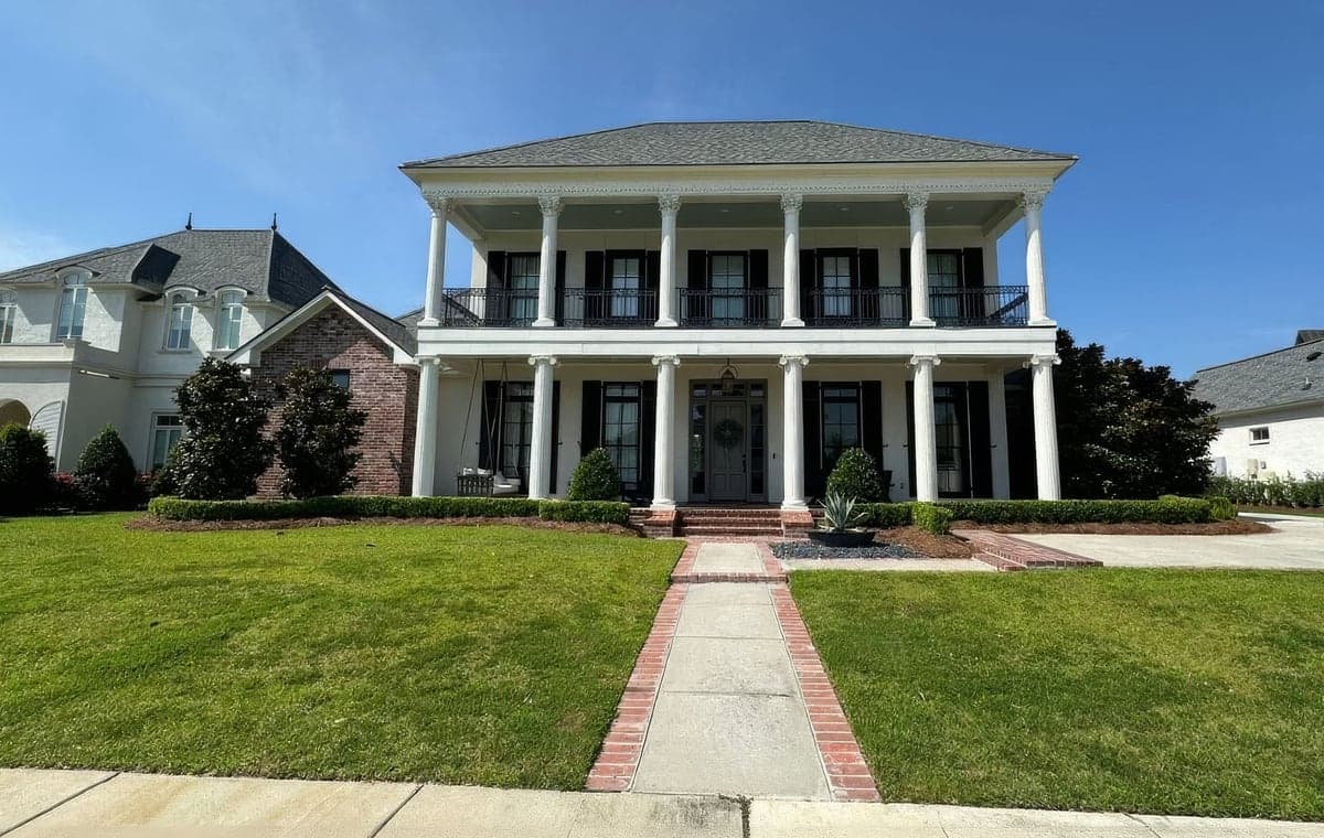 A two-story, white columned Southern-style home with black shutters and a second-floor balcony, fronted by a neatly manicured green lawn, trimmed boxwood hedges, and a brick-edged walkway. The landscaping appears freshly