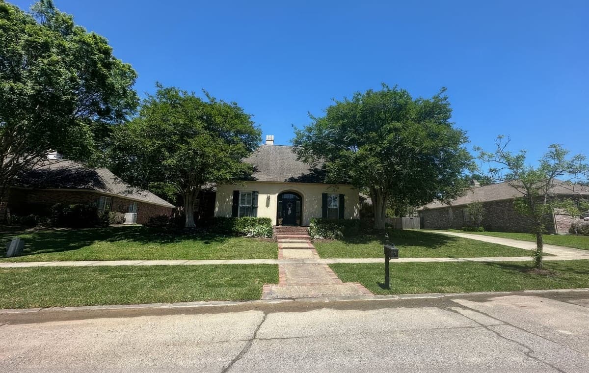 A well-maintained residential front yard with a neatly mowed green lawn, trimmed shrubs along the foundation, and mature shade trees in front of a cream stucco home with a brick walkway. The clear blue sky and crisp lawn