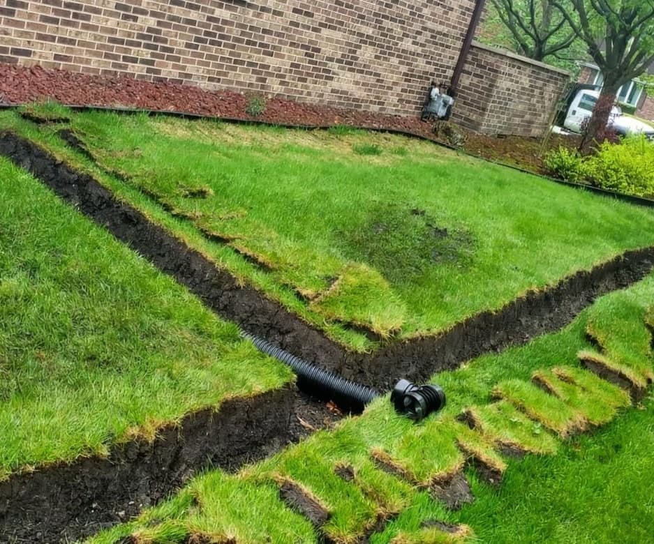 A trench dug through a front lawn beside a brick home with sod strips peeled back and corrugated black drainage pipe and a fitting laid out, showing a yard drainage system installation in progress