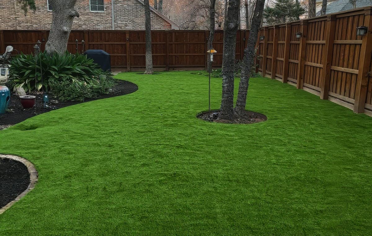 Artificial turf in a fenced backyard with curved mulch beds and mature trees