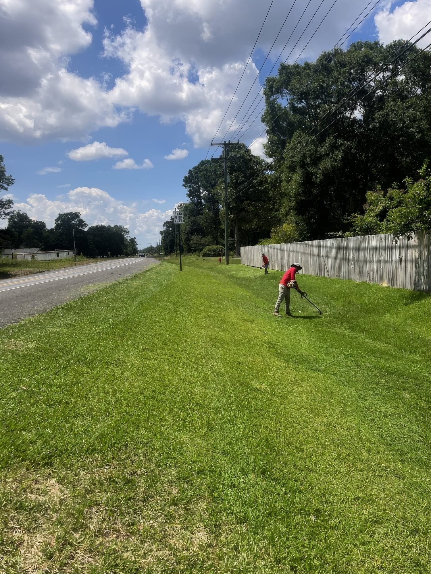 Two crew members trimming a long grassy roadside embankment