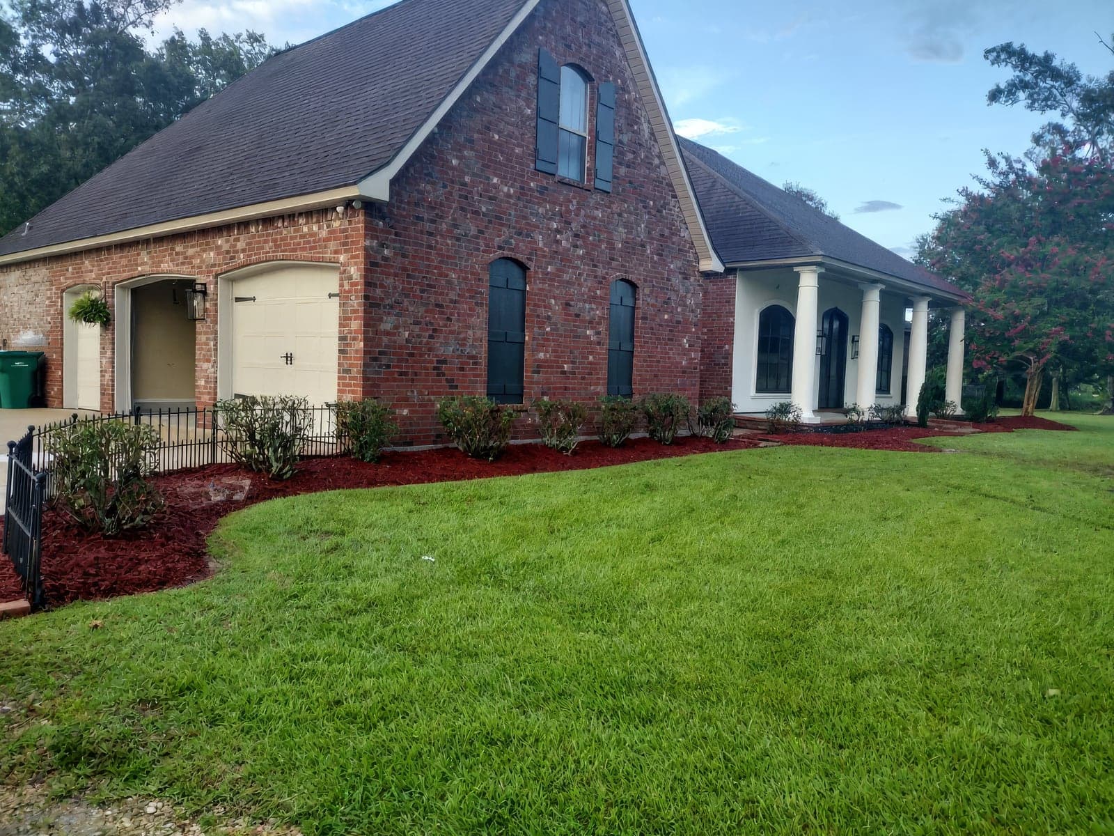 A red brick home with a manicured green lawn and freshly installed red mulch beds lined with neatly trimmed shrubs along the foundation and wrought iron fence. The completed landscaping frames the house's white columned