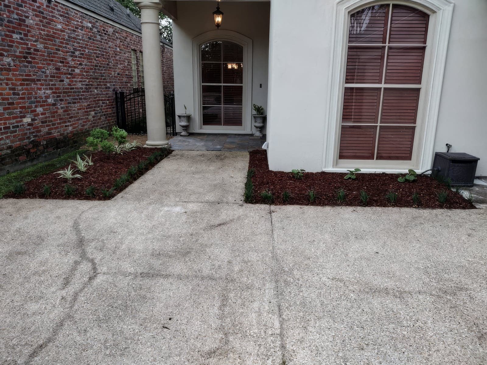 Freshly installed landscaping beds with dark red mulch and young plants flank a concrete walkway leading to the covered front entry of a stucco home with brick neighboring wall. The newly completed beds show clean edges