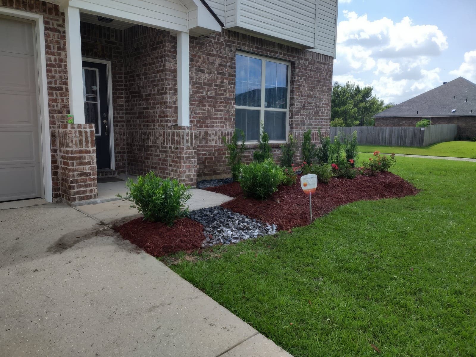 Freshly installed landscaping bed in front of a brick home, featuring red mulch, decorative gray river rock, evergreen shrubs, and blooming red rose bushes along a clean lawn edge