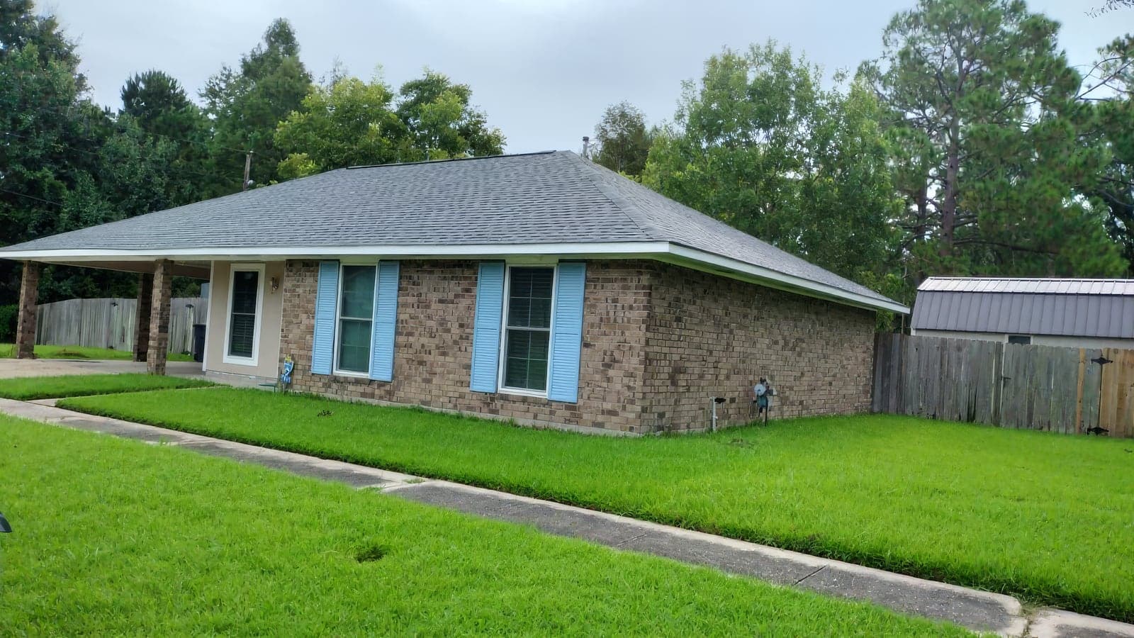 Single-story brick ranch home with blue shutters, a carport, and a freshly maintained, vibrant green lawn with trimmed edges along the sidewalk. A wooden privacy fence and storage shed are visible at the side of the prop