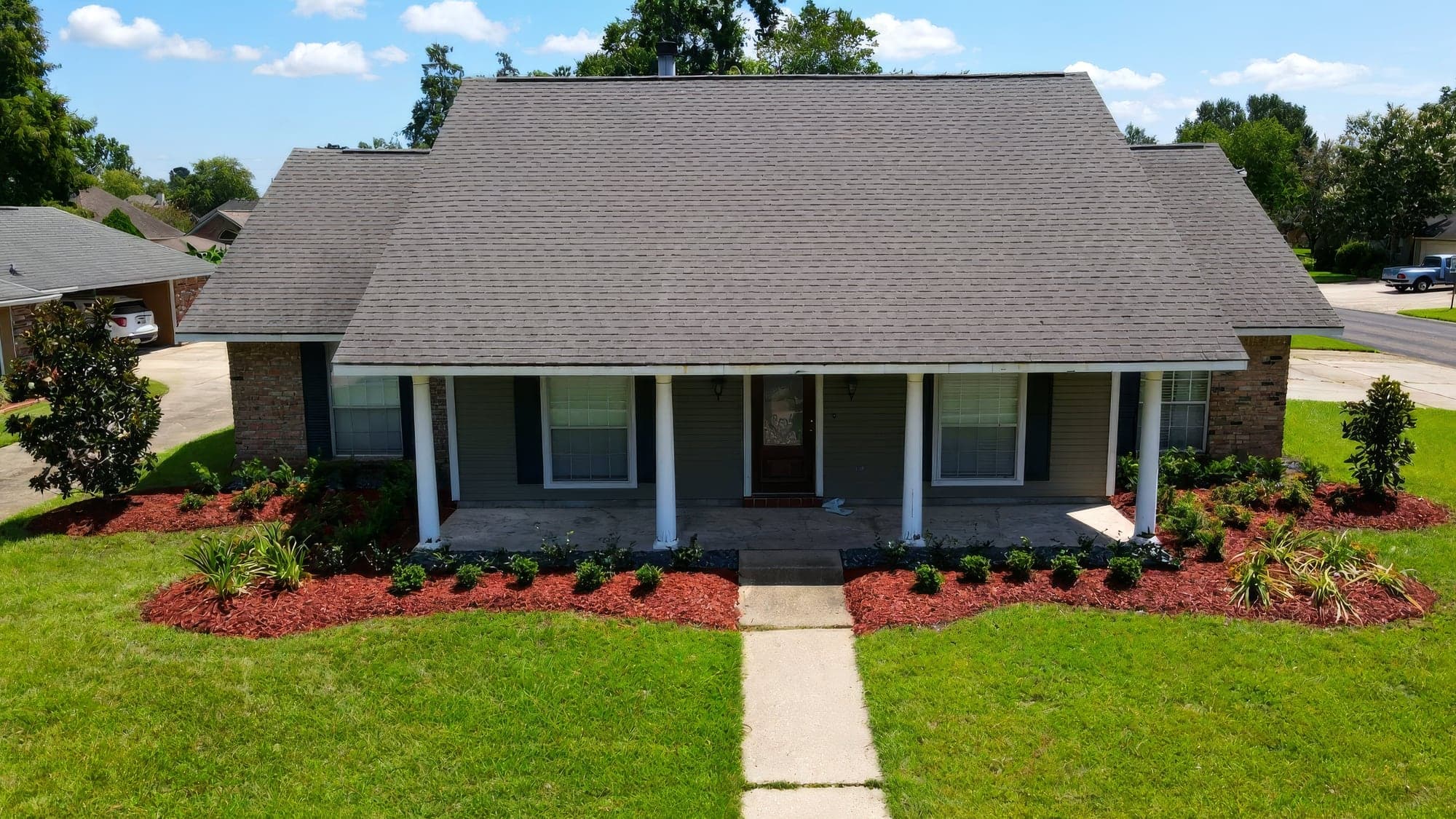 Aerial view of a single-story brick ranch home with a covered front porch, featuring freshly installed landscaping with red mulch beds, young shrubs, ornamental grasses, and a neatly manicured lawn