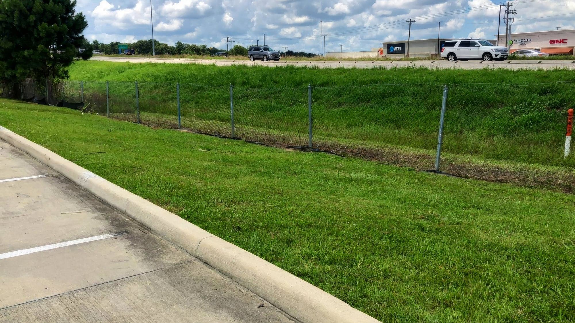 A manicured grass embankment beside a concrete curb and parking area, with a chain-link fence at the base of the slope and a busy road and strip-mall storefronts (AT&T, GNC) in the background