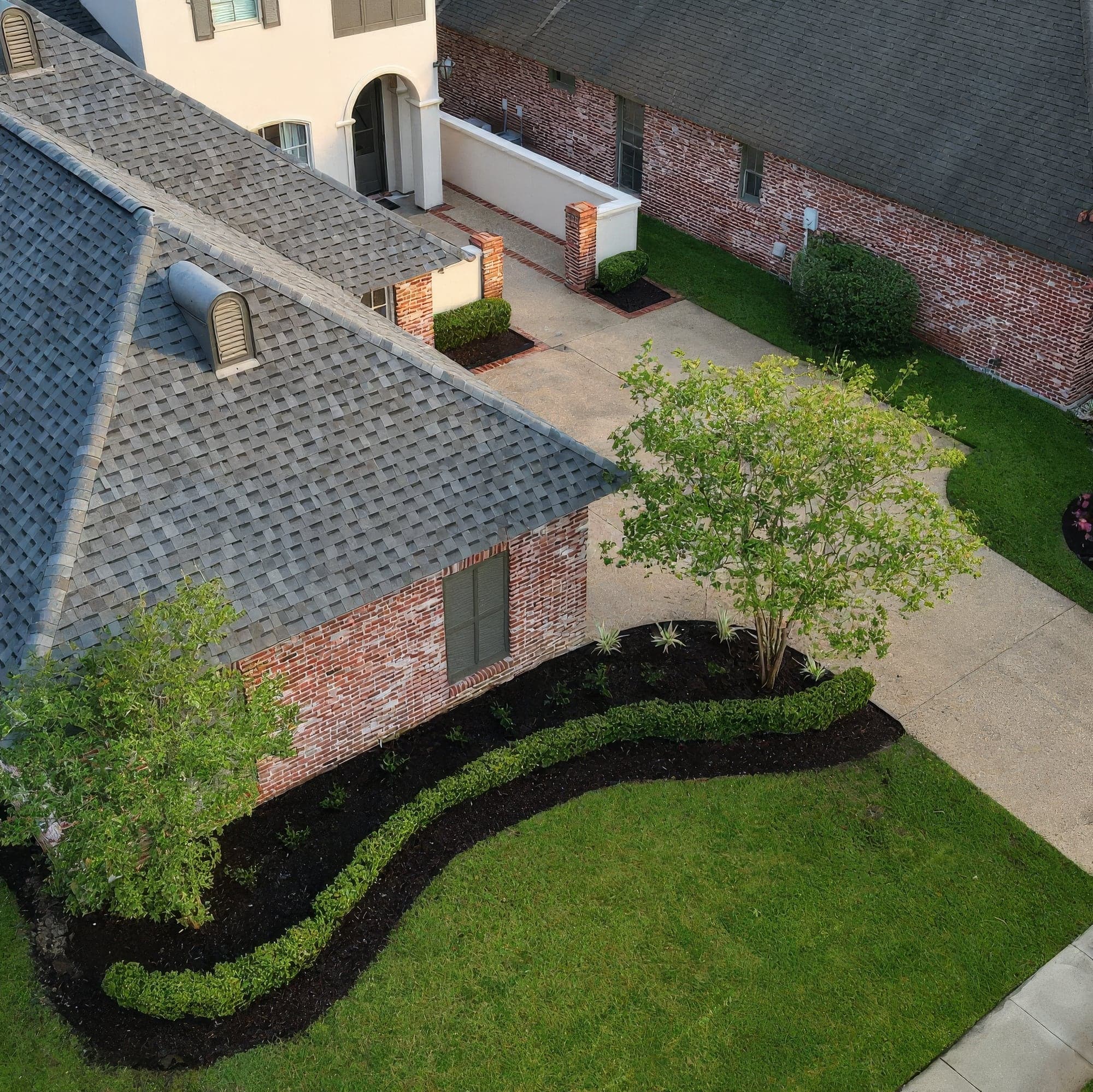 Aerial view of a freshly landscaped brick home with a curved bed of dark mulch, a neatly trimmed boxwood hedge border, young crape myrtle tree, and new plantings alongside a manicured green lawn