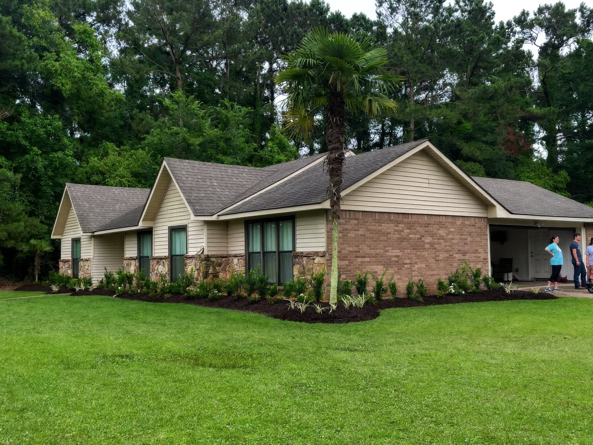 A single-story ranch-style home with a freshly landscaped front bed featuring dark mulch, young shrubs, flowering plants, and a tall windmill palm, set against a lush green lawn and wooded backdrop. A few people stand ne