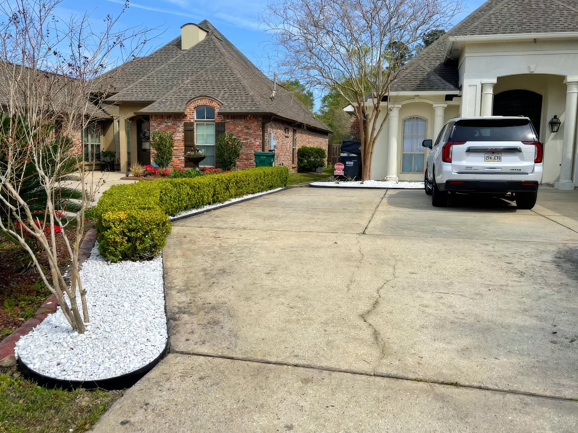 Freshly installed white decorative rock landscape beds with black edging border a trimmed boxwood hedge along a residential concrete driveway. Colorful azaleas and a well-kept brick home are visible in the background
