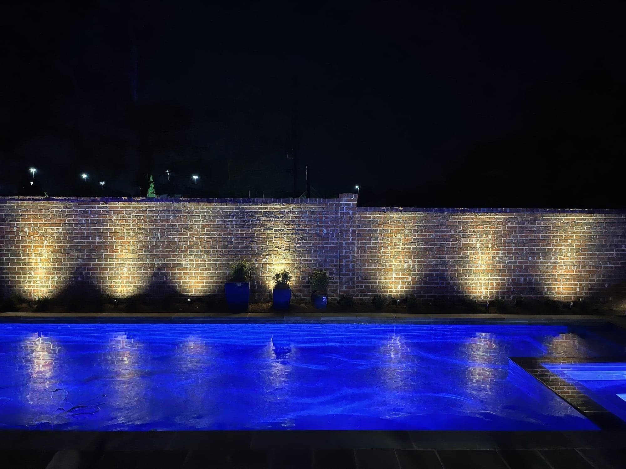 Low-voltage landscape lights washing a brick wall beside a pool at night
