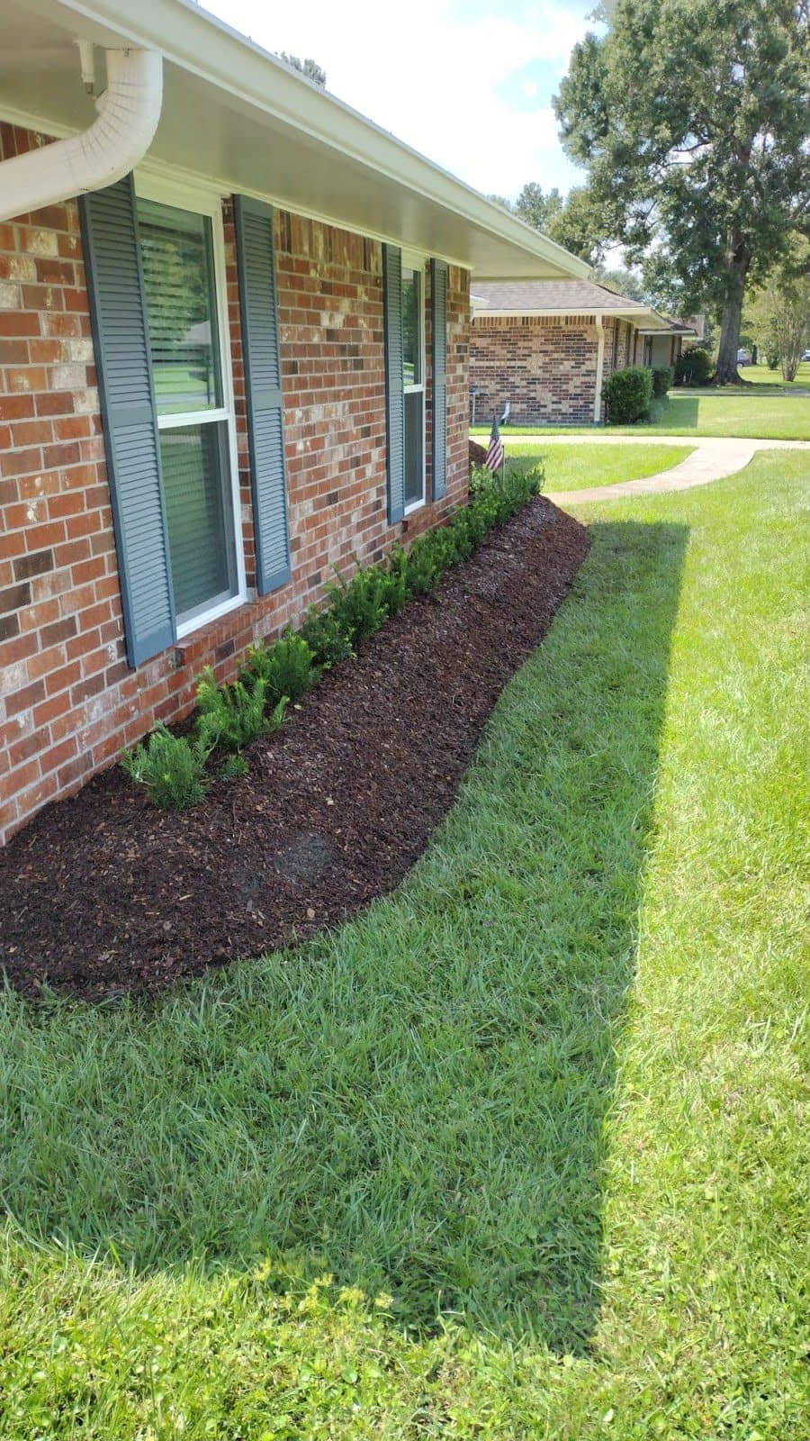A freshly mulched garden bed with newly planted shrubs lines the brick exterior of a home, showcasing completed landscaping work