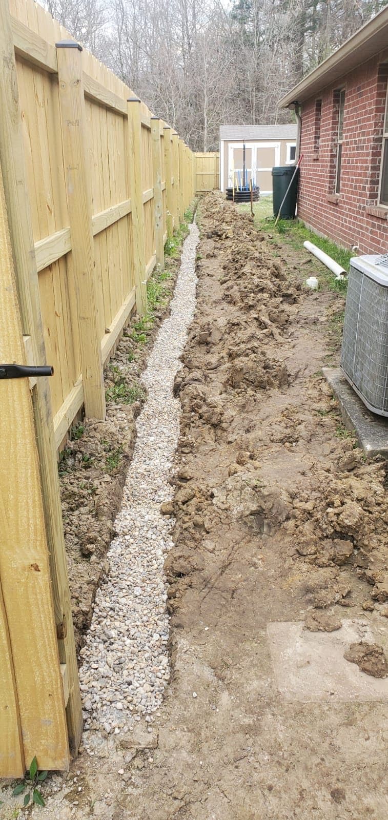 A French drain trench filled with washed gravel along a wooden privacy fence