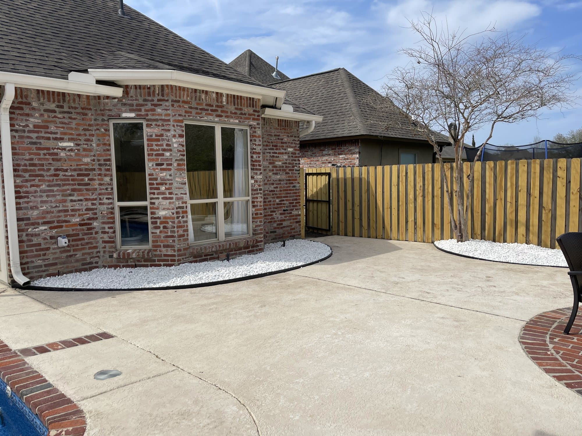 Freshly completed landscaping beside a brick home and pool patio, featuring curved black edging filled with bright white decorative rock along the house and around a crepe myrtle tree, next to a new wood privacy fence an
