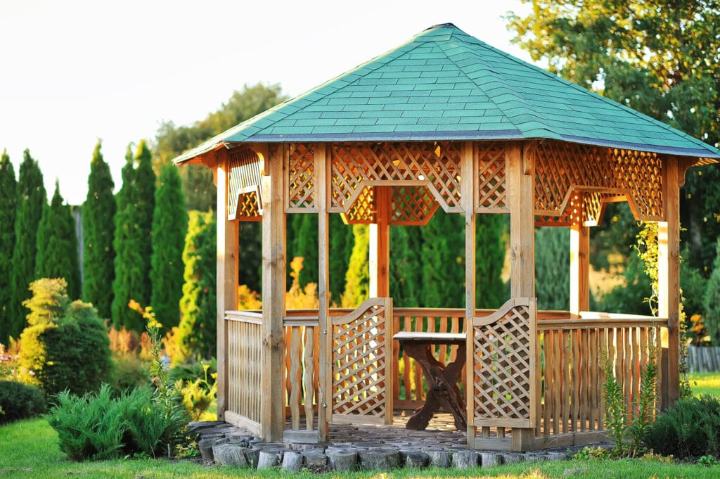A wooden gazebo with a green tile roof amid a lush green landscape bathed in the light of the sunset.