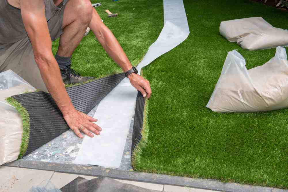 A person pulling up and replacing vibrant green artificial turf with bags of sand and tools in the background.