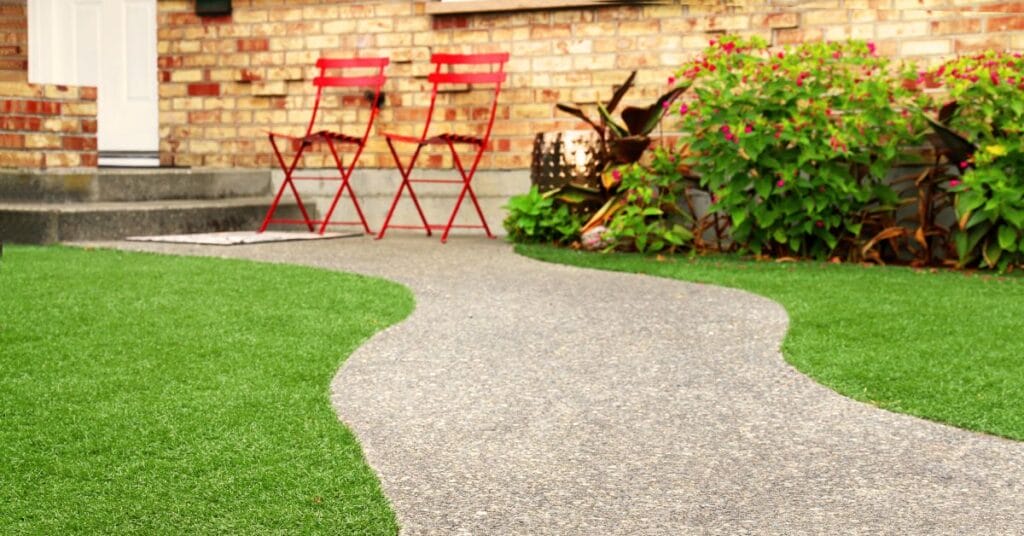 A back garden with a short, gray pathway cutting through artificial grass toward a set of red chairs.