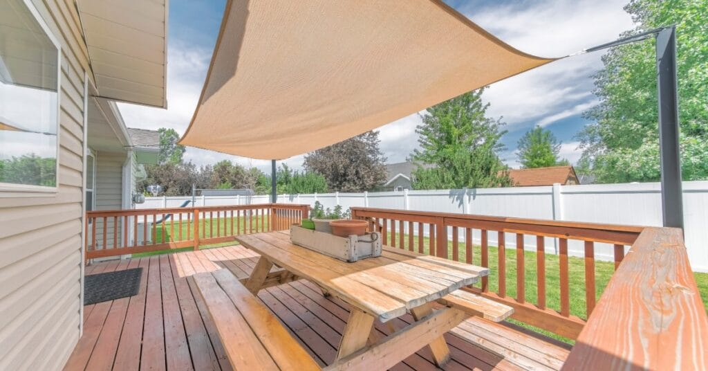 A small backyard with a wooden patio and a wooden picnic table under a beige shade sail during a bright, sunny day.