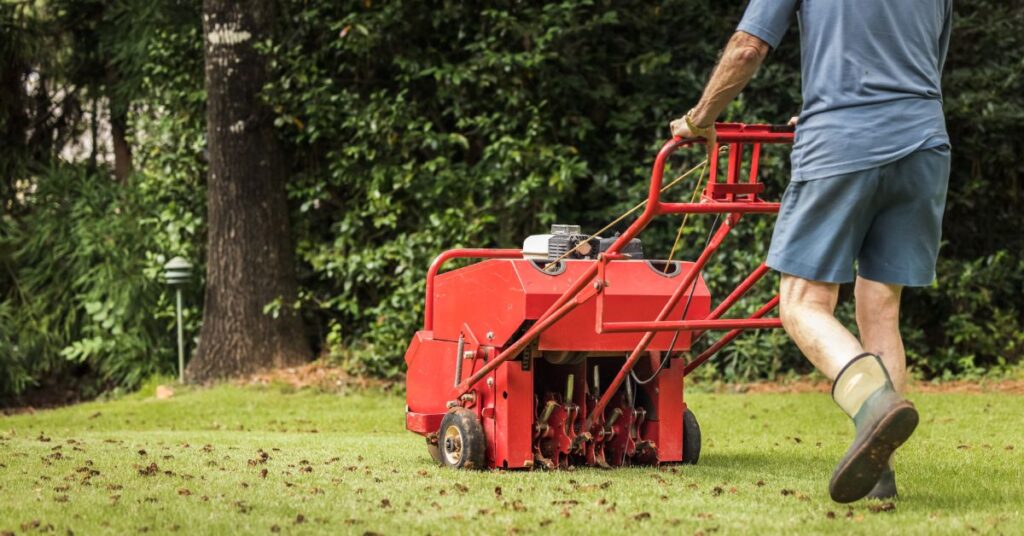 A man wearing gloves and boots pushing a red, gas-powered aerating machine across his residential lawn.